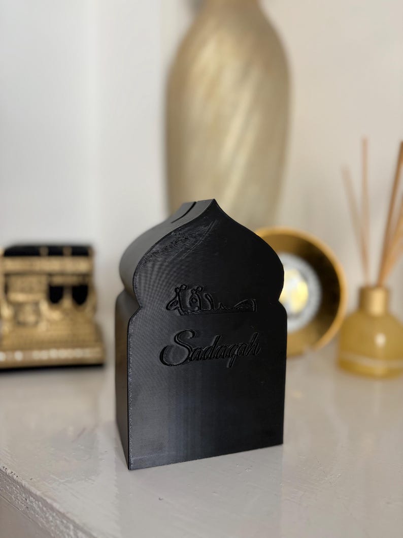 Puede incluir: Caja decorativa negra con una parte superior arqueada y la palabra "Sadaqah" grabada en la parte delantera. La caja tiene un dise&ntilde;o similar a una mezquita y probablemente se utiliza para donaciones ben&eacute;ficas. Otros elementos decorativos est&aacute;n en el fondo.