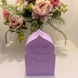 Puede incluir: Una caja Sadagah de color lila claro con una parte superior arqueada, con las palabras "Qudsiyya", "Sadagah" en escritura &aacute;rabe y "Sadagah" en ingl&eacute;s. Un ramo de flores rosas y melocot&oacute;n est&aacute; en el fondo.