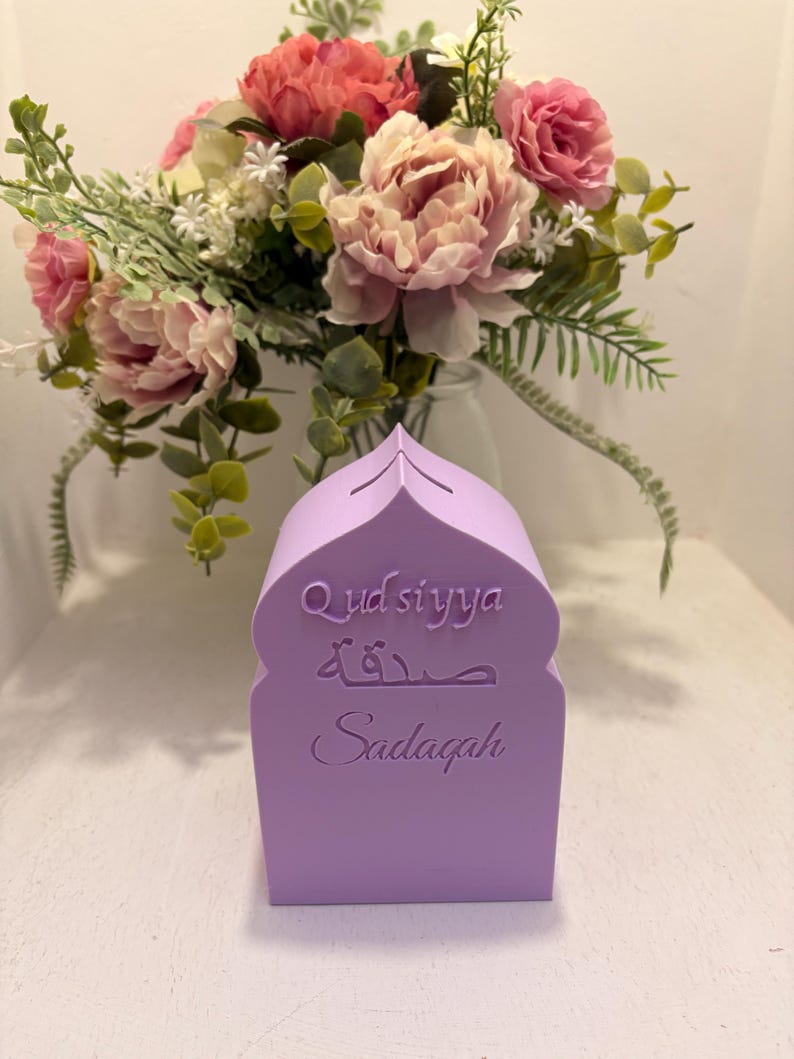 Puede incluir: Una caja Sadagah de color lila claro con una parte superior arqueada, con las palabras "Qudsiyya", "Sadagah" en escritura &aacute;rabe y "Sadagah" en ingl&eacute;s. Un ramo de flores rosas y melocot&oacute;n est&aacute; en el fondo.