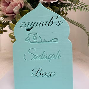 Puede incluir: Una caja Sadagah azul claro con una parte superior arqueada. La caja tiene el nombre "Zaynab's" y la palabra "Sadagah" en ingl&eacute;s y &aacute;rabe. Las palabras "Sadagah Box" tambi&eacute;n est&aacute;n grabadas.
