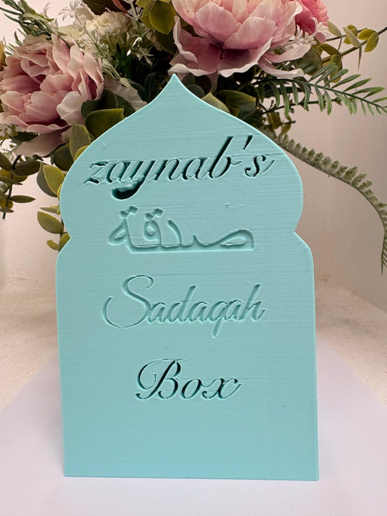 Puede incluir: Una caja Sadagah azul claro con una parte superior arqueada. La caja tiene el nombre "Zaynab's" y la palabra "Sadagah" en ingl&eacute;s y &aacute;rabe. Las palabras "Sadagah Box" tambi&eacute;n est&aacute;n grabadas.