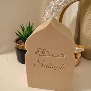Puede incluir: Una caja Sadaqah beige con forma de mezquita con escritura &aacute;rabe grabada y la palabra "Sadaqah" debajo. Una peque&ntilde;a planta en maceta y una luna creciente decorativa est&aacute;n en el fondo. La caja es de material claro.