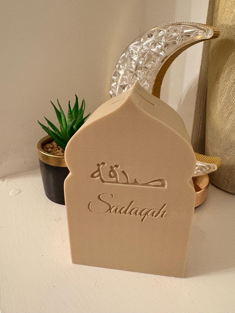 Puede incluir: Una caja Sadaqah beige con forma de mezquita con escritura &aacute;rabe grabada y la palabra "Sadaqah" debajo. Una peque&ntilde;a planta en maceta y una luna creciente decorativa est&aacute;n en el fondo. La caja es de material claro.