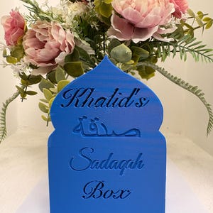 Puede incluir: Una caja Sadagah azul con el nombre "Khalid's" y las palabras "Sadagah Box" en ingl&eacute;s y &aacute;rabe. La caja est&aacute; frente a un ramo de flores rosas y melocot&oacute;n con follaje verde.