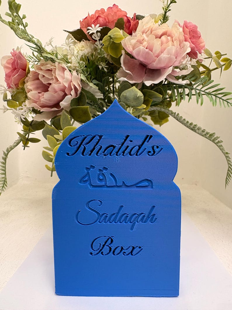 Puede incluir: Una caja Sadagah azul con el nombre "Khalid's" y las palabras "Sadagah Box" en ingl&eacute;s y &aacute;rabe. La caja est&aacute; frente a un ramo de flores rosas y melocot&oacute;n con follaje verde.