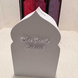 Puede incluir: Una hucha blanca con forma de mezquita con el texto "Khan Family Sadaqah" en relieve en la parte delantera. El fondo presenta dos faroles decorativos con paneles de vidrieras rojas y moradas.