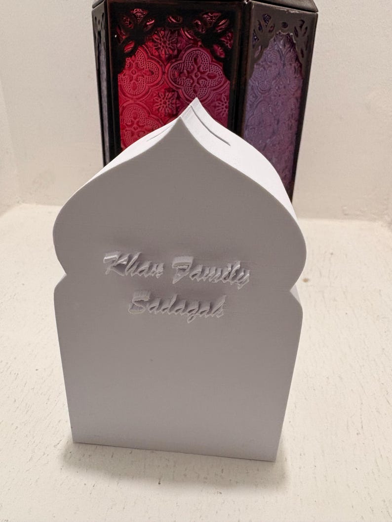 Puede incluir: Una hucha blanca con forma de mezquita con el texto "Khan Family Sadaqah" en relieve en la parte delantera. El fondo presenta dos faroles decorativos con paneles de vidrieras rojas y moradas.