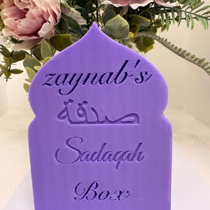 Puede incluir: Una caja de Sadaqah morada con el nombre "zaynab's" y escritura &aacute;rabe grabada en la parte delantera. La caja tiene una parte superior decorativa en forma de arco y la palabra "Box" en la parte inferior. El fondo presenta un arreglo floral con flores rosas y verdes.