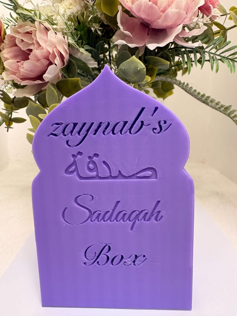 Puede incluir: Una caja de Sadaqah morada con el nombre "zaynab's" y escritura &aacute;rabe grabada en la parte delantera. La caja tiene una parte superior decorativa en forma de arco y la palabra "Box" en la parte inferior. El fondo presenta un arreglo floral con flores rosas y verdes.