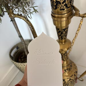 Puede incluir: Una tarjeta blanca con una parte superior arqueada, con escritura &aacute;rabe y la palabra "Sadaqah" en cursiva. La tarjeta se sostiene frente a una cachimba decorativa de lat&oacute;n y una peque&ntilde;a planta en maceta.
