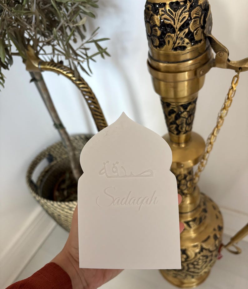 Puede incluir: Una tarjeta blanca con una parte superior arqueada, con escritura &aacute;rabe y la palabra "Sadaqah" en cursiva. La tarjeta se sostiene frente a una cachimba decorativa de lat&oacute;n y una peque&ntilde;a planta en maceta.