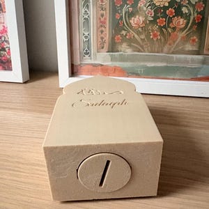 Puede incluir: Una hucha rectangular beige con una ranura para monedas. El banco tiene una parte superior arqueada con escritura &aacute;rabe grabada. El banco est&aacute; sobre una superficie de madera. El fondo incluye obras de arte enmarcadas con dise&ntilde;os florales.