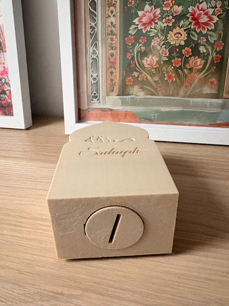 Puede incluir: Una hucha rectangular beige con una ranura para monedas. El banco tiene una parte superior arqueada con escritura &aacute;rabe grabada. El banco est&aacute; sobre una superficie de madera. El fondo incluye obras de arte enmarcadas con dise&ntilde;os florales.