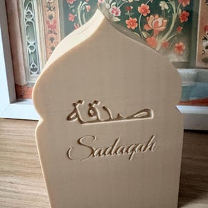Puede incluir: Una pieza decorativa beige, con forma de arco, con la palabra "Sadaqah" en ingl&eacute;s y escritura &aacute;rabe. La pieza est&aacute; hecha de un material de color claro y se apoya sobre una superficie de madera, con una pintura floral en el fondo.