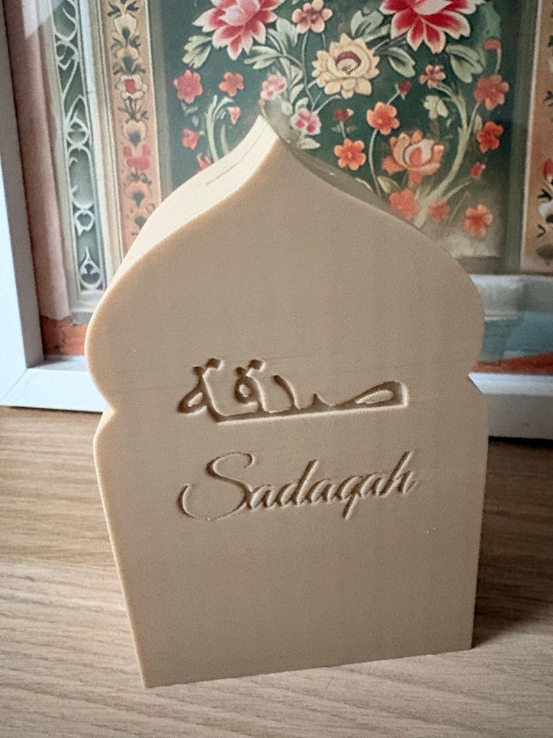 Puede incluir: Una pieza decorativa beige, con forma de arco, con la palabra "Sadaqah" en ingl&eacute;s y escritura &aacute;rabe. La pieza est&aacute; hecha de un material de color claro y se apoya sobre una superficie de madera, con una pintura floral en el fondo.