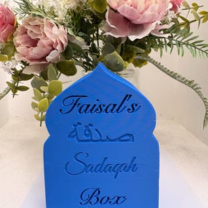 Puede incluir: Una caja Sadaqah azul con el nombre "Faisal's" y las palabras "Sadaqah Box" grabadas en la parte delantera. La caja tiene una parte superior arqueada y est&aacute; frente a un ramo de flores rosas y blancas.