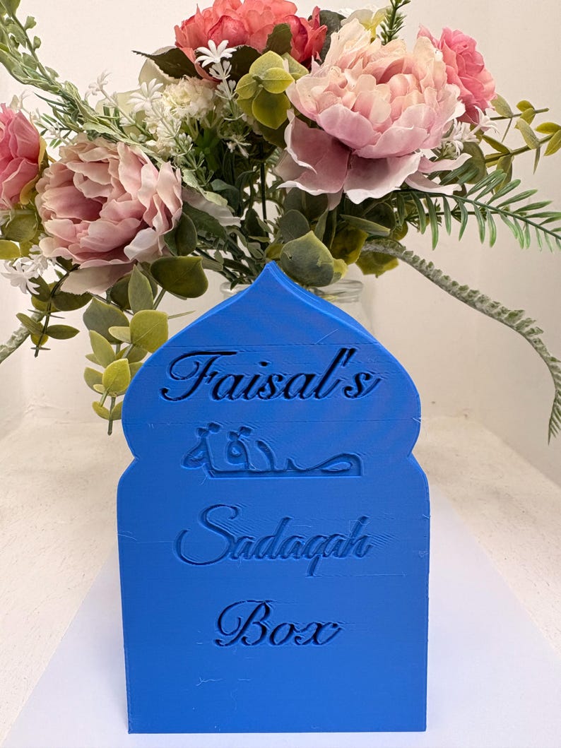 Puede incluir: Una caja Sadaqah azul con el nombre "Faisal's" y las palabras "Sadaqah Box" grabadas en la parte delantera. La caja tiene una parte superior arqueada y est&aacute; frente a un ramo de flores rosas y blancas.