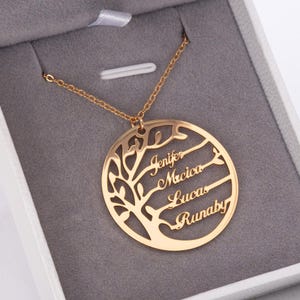 Puede incluir: Un collar de árbol genealógico dorado con los nombres Jenifer, Mrcica, Lucas y Runaby grabados. El colgante es circular con un diseño de árbol y cuelga de una delicada cadena. El collar se presenta en una caja de joyería gris.