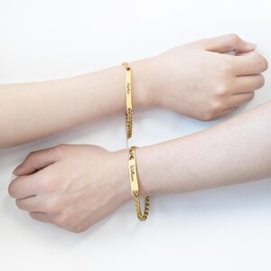 Puede incluir: Dos pulseras doradas con eslabones de cadena y placas rectangulares con nombres. Una pulsera dice "Sophia" y la otra "William". Las pulseras se muestran en dos muñecas sobre un fondo blanco.