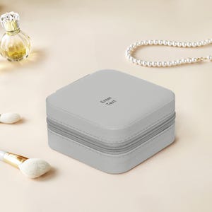 Puede incluir: Una caja de joyería cuadrada de color gris claro con cierre de cremallera. El texto "Enter Text" está impreso en la parte superior. Un frasco de perfume, brochas de maquillaje y un collar de perlas están en el fondo.