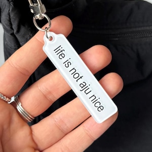 May include: A white rectangular keyring with the phrase "life is not aju nice" printed in black. The keyring is attached to a silver metal clasp and a black bag.