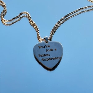 Può includere: Una collana con ciondolo a forma di cuore argentato con una catena dorata. Il ciondolo reca l'incisione "You're just a Fallen Superstar". La collana è esposta su una superficie riflettente.