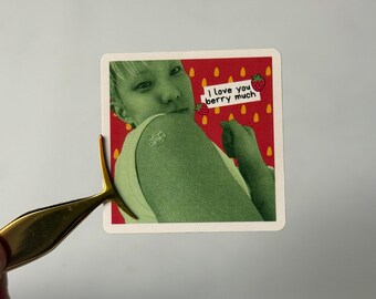 Può includere: Adesivo quadrato con un'immagine in tonalità verde di una persona. L'adesivo ha uno sfondo rosso con testo bianco che recita "I love you berry much" e illustrazioni di fragole. L'adesivo è tenuto da una pinzetta dorata.