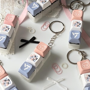 Peut inclure: Porte-clés avec des touches de clavier empilées aux couleurs pastel : rose, blanc et bleu. Chaque porte-clés est orné d'un anneau argenté et d'un motif avec du texte et un personnage de dessin animé. Rubans et trombones sont également présents.