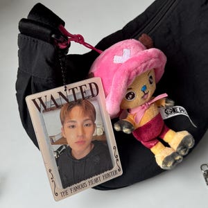 Op de afbeelding: Zwarte nylon heuptas met een roze rits en een klein pluche speelgoed van een stripfiguur met een roze hoed. Een doorzichtige plastic kaart met een foto en de tekst "WANTED" is aan de rits bevestigd.