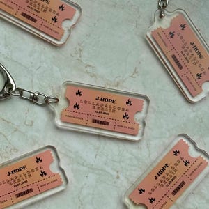 Puede incluir: Llaveros de acrílico transparente con diseño de entrada. El texto en la entrada dice "J-HOPE LOLLAPALOOZA BERLIN 13.07.2023". Los llaveros tienen un cierre y un anillo de metal plateado. El diseño de la entrada es melocotón y naranja.