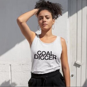 May include: A light gray sleeveless tank top featuring the text GOAL DIGGER printed in bold black capital letters across the chest. The garment has a simple round neckline and is paired with black trousers that have a gathered, elasticized waist.