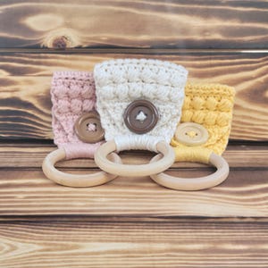 May include: Three crocheted teethers with wooden rings. The teethers are in pink, white, and yellow with brown buttons.