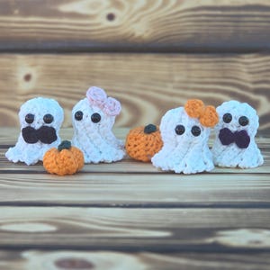 Può includere: Una collezione di decorazioni di Halloween fatte a mano all'uncinetto. L'immagine presenta figure di fantasmi bianchi con occhi a bottone neri e papillon o fiocchi, insieme a piccole zucche arancioni. Gli oggetti sono disposti su una superficie di legno.