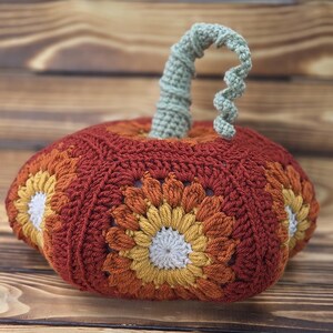 May include: A handmade crochet pumpkin in shades of orange, rust, and yellow, with a light green stem. The pumpkin features a floral design on its sides and is set against a wooden background. A decorative item for fall.
