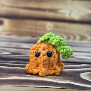 Puede incluir: Una criatura de ganchillo naranja hecha a mano con una parte superior verde y dos ojos de seguridad negros. El amigurumi está sentado sobre una superficie de madera. El juguete de ganchillo es un diseño único y caprichoso.