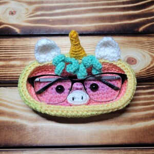May include: A crocheted unicorn-shaped glasses holder with a pink, yellow, and green color scheme. The holder has a pair of glasses inside and a yellow horn on top.
