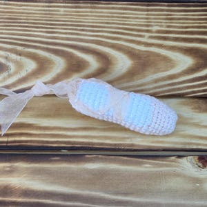 May include: A handmade, crocheted ballet slipper in white and light blue, tied with a sheer ribbon. The slipper is lying on a wooden surface with a natural wood grain pattern. The overall aesthetic is soft and delicate.