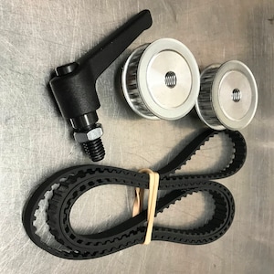 May include: A black rubber timing belt with a silver metal tensioner and a black plastic lever. The belt is folded in half and secured with a rubber band.