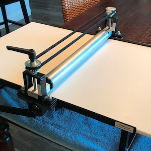 Peut inclure: Une presse d'impression de table blanche avec un cadre en métal noir et un rouleau argenté. La presse a une poignée en bois et une courroie noire. La presse est étiquetée "JT Designs".