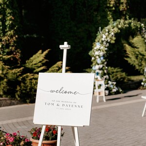 Op de afbeelding: Een witte ezel met een bord waarop staat "welcome to the wedding of Tom & Dayenne 13 July 2024". De ezel staat op een betegeld terras met groene planten en bloemen in potten.