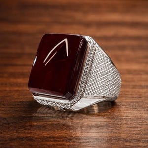 Pode incluir: Um anel de prata com uma pedra grande, retangular e vermelho escuro. A pedra tem um design branco. A banda do anel é incrustada com pequenas pedras transparentes. O anel é exibido em uma superfície de madeira.