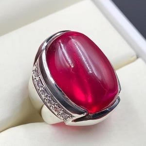 May include: A silver ring featuring a large, oval-shaped, translucent red gemstone. The ring has a polished silver band and a row of small, clear stones set along the side of the gemstone. The gemstone is the focal point.
