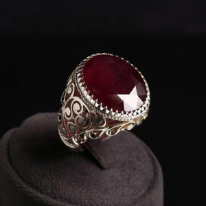 May include: A silver ring featuring a large, oval-cut, deep red gemstone. The ring's band has an intricate, openwork pattern, and the gemstone is framed by a serrated edge. The ring is presented on a dark grey surface against a black backdrop.