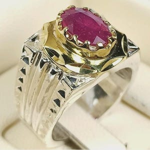 May include: A two-tone ring with a large, oval-cut ruby gemstone. The ring has a silver band with a textured design and a gold-coloured setting around the ruby. The ruby is a deep red colour.