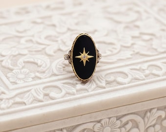 May include: An oval-shaped ring with a black onyx center featuring a gold starburst design. The ring has a gold band with ornate detailing and small accent stones on each side. The ring is set on a white, carved surface.