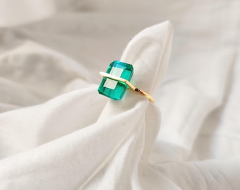 Op de afbeelding: Een gouden ring met een grote, smaragdgeslepen, transparante groene edelsteen. De band en zetting van de ring zijn goudkleurig. De ring wordt gepresenteerd op een witte stoffen achtergrond.