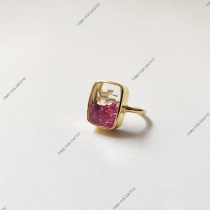 Puede incluir: Un anillo dorado con un engaste cuadrado y transparente lleno de pequeñas piedras preciosas rosas vibrantes. El anillo tiene una banda delicada. El texto "TIME FOR EXOTIC" es visible en el fondo.
