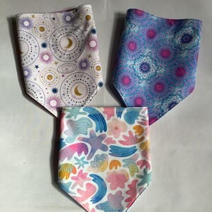 May include: Three fabric bandanas with different patterns. One bandana has a white background with a purple, gold, and blue pattern of stars, moons, and circles. Another bandana has a blue background with a pink and purple pattern of geometric shapes. The third bandana has a white background with a pink, blue, and yellow pattern of abstract shapes.