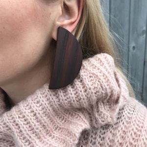 May include: A pair of large, dark brown wooden earrings in the shape of a half-circle. The earrings are worn by a person with blonde hair and a pink sweater.