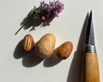 May include: Three small wooden eggs, one with a striped pattern, and a small purple flower on a white surface. A wooden handled carving knife with a silver blade is also in the image.